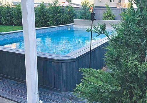 A residential backyard with a rectangular above-ground swimming pool surrounded by wooden decking, and greenery.